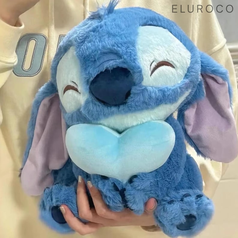 May include: A plush blue Stitch character with large ears and a heart-shaped chest. The soft toy features a two-tone design, with a lighter blue face and a darker blue body. The character's eyes are closed in a peaceful expression.