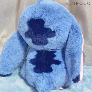 May include: A plush toy in shades of blue, featuring a distinctive dark blue patch on its back. The toy has large ears and a soft, furry texture. The text "ELUROCO" is visible in the upper right corner. The toy is a character from a popular animated film.