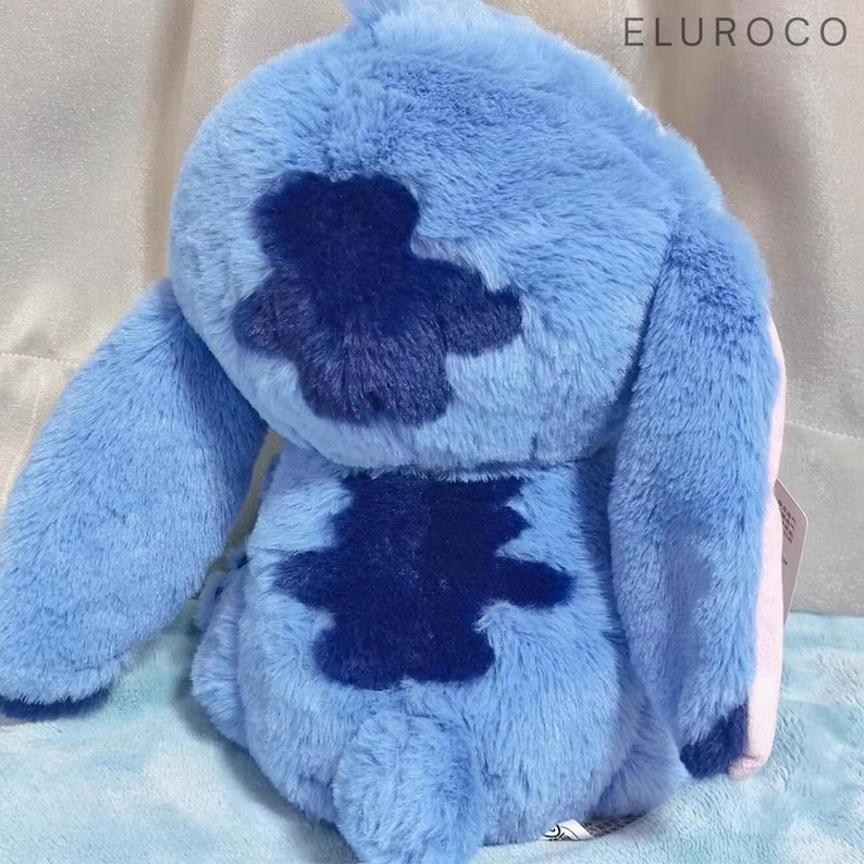 May include: A plush toy in shades of blue, featuring a distinctive dark blue patch on its back. The toy has large ears and a soft, furry texture. The text "ELUROCO" is visible in the upper right corner. The toy is a character from a popular animated film.