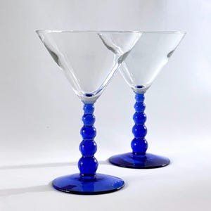 May include: Two clear martini glasses with vibrant blue stems and bases. The stems feature a series of spherical accents, adding a unique design element. The glasses are set against a plain white background, highlighting their elegant form.