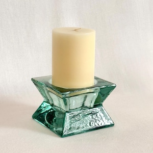 May include: A light beige pillar candle sits in a square, teal-colored glass candle holder. The candle holder has a geometric design with a textured surface. The background is a plain white fabric.