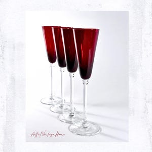 May include: Four tall champagne flutes with deep red glass bowls and clear glass stems and bases. The flutes are arranged in a staggered row, showcasing the vibrant crimson color of the glass against a plain white background. The text Artful Vintage Home is visible in the lower left corner.