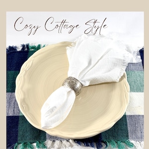 May include: A cream-colored ceramic plate with a scalloped edge, a white fabric napkin with frayed edges, and a silver-toned metal napkin ring with an embossed floral pattern. The text "Cozy Cottage Style" is visible at the top of the image.