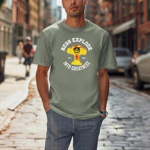 May include: Sage green t-shirt with a graphic that reads "REGS EXPLODE INTO GREATNESS" in a circular design. The graphic features a yellow explosion with a lion's head in the center. The text "EST. 1990" is also included.