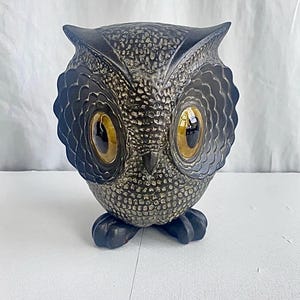 May include: A ceramic owl figurine featuring a textured, speckled grey and black body with large, glossy, amber-colored eyes. The owl has prominent, scalloped facial discs surrounding the eyes and dark, solid-colored feet at the base.