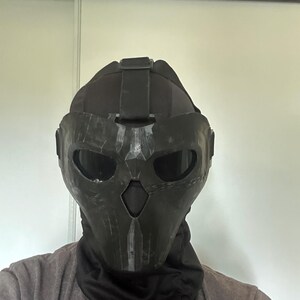Puede incluir: Una m&aacute;scara t&aacute;ctica negra con un dise&ntilde;o de calavera, con aberturas para los ojos y la boca. La m&aacute;scara se usa con un cuello negro y una camiseta gris. La m&aacute;scara parece estar hecha de un material duro y duradero.