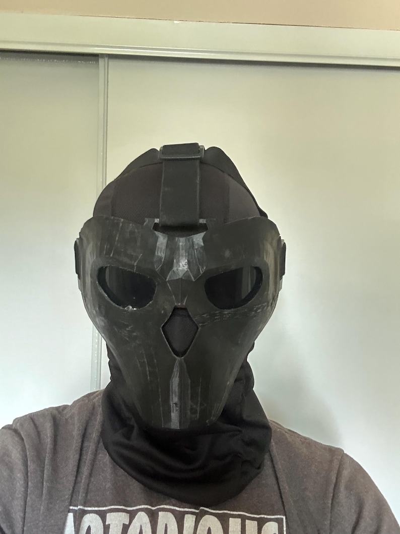 Puede incluir: Una m&aacute;scara t&aacute;ctica negra con un dise&ntilde;o de calavera, con aberturas para los ojos y la boca. La m&aacute;scara se usa con un cuello negro y una camiseta gris. La m&aacute;scara parece estar hecha de un material duro y duradero.