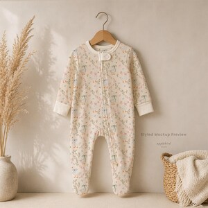May include: A long-sleeved, footed baby sleeper in a cream color featuring a repeating pattern of white geese with green bows, green leaves, and small pink flowers. The garment includes a front zipper closure with a snap tab at the neckline.