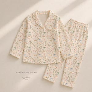 May include: A two-piece pajama set in a cream color featuring a repeating pattern of white geese with blue and green bows, interspersed with small pink flowers and green foliage. The long-sleeve button-down shirt has a collar and a chest pocket.