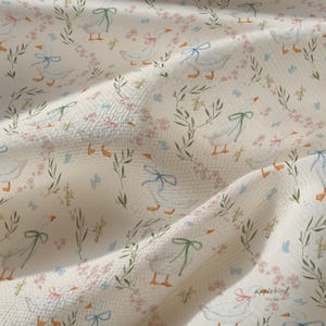 May include: A white fabric with a textured waffle weave pattern featuring a repeating print of white geese with orange beaks and feet. The geese are adorned with small pink, blue, or green bows around their necks, surrounded by green foliage.