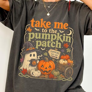 Pode incluir: Uma t-shirt cinzento-carvão de manga curta com uma impressão gráfica com o texto "take me to the pumpkin patch". O design inclui um fantasma com um chapéu de bruxa, uma abóbora, uma placa de madeira "PUMPKINS THIS WAY" e um carrinho.