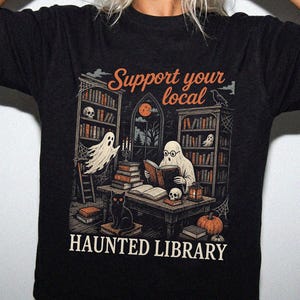 Könnte beinhalten: Ein schwarzes T-Shirt mit einem Grafikdruck einer Bibliotheksszene mit Geistern, einer schwarzen Katze und einem Kürbis. Der Text lautet Support your local HAUNTED LIBRARY. Die Illustration zeigt Bücherregale, einen Schreibtisch und einen Totenkopf in Orange-, Weiß- und Grautönen auf dem dunklen Stoff.
