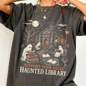 Op de afbeelding: Een antracietgrijs t-shirt met een afbeelding van een spookbibliotheek met geesten die boeken lezen, een kale boom en een volle maan. De tekst luidt SUPPORT YOUR LOCAL HAUNTED LIBRARY. De afbeelding bevat stapels boeken en een zwarte kat.
