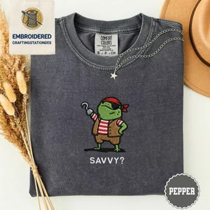 Könnte beinhalten: Dunkelgraues T-Shirt mit einer Stickerei einer Piratenfrosch-Design. Der Frosch trägt ein rotes Bandana, eine Augenklappe und eine braune Weste. Der Text "SAVVY?" steht darunter. Eine silberne Halskette mit Sternanhänger liegt auf dem Shirt.
