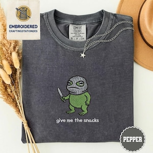May include: Dark gray t-shirt with an embroidered green frog wearing a ski mask and holding a knife. The text "give me the snacks" is below the frog. A silver necklace with a star pendant is on the shirt.