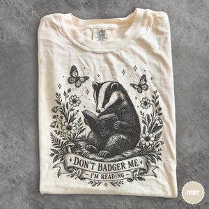 May include: An ivory t-shirt featuring a black ink illustration of a badger reading a book, surrounded by flowers, butterflies, and stars. A banner at the bottom contains the text, "DON'T BADGER ME - I'M READING." The shirt has a crew neckline.