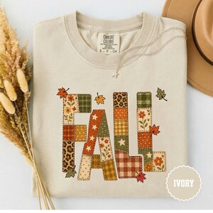 May include: An ivory short-sleeve t-shirt featuring the word FALL in large block letters. Each letter is composed of various patchwork patterns including leopard print, plaid, stars, and floral designs in orange, green, and brown tones.