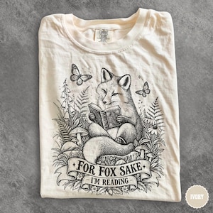May include: An ivory t-shirt featuring a black ink illustration of a fox reading a book while sitting on a stump. The design includes ferns, mushrooms, flowers, and butterflies. A banner at the bottom contains the text, "FOR FOX SAKE I'M READING."