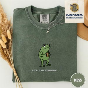 Op de afbeelding: Een mosgroen t-shirt met een cartoon kikker die een koffiekopje vasthoudt. De tekst "PEOPLE ARE EXHAUSTING" staat eronder. Een zilveren sterrenketting en een label met "EMBROIDERED CRAFTINGSTATIONDES" zijn ook zichtbaar.