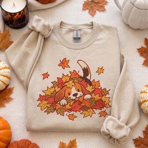 Peut inclure: Un sweat-shirt à col rond beige avec une illustration graphique d'un chiot beagle marron et blanc jouant dans un tas de feuilles d'automne colorées. Le motif comprend des feuilles d'érable orange, jaunes et rouges autour du chiot.