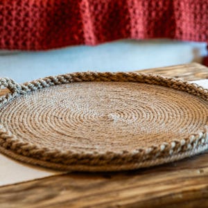 May include: A round tray constructed from coiled natural tan jute rope. The tray features a flat circular base with a raised, braided rope border and two integrated rope handles on opposite sides, creating a textured, circular serving surface.