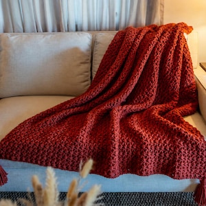 May include: A chunky knit throw blanket in a deep burnt orange colour, featuring a textured waffle-like stitch pattern throughout the fabric. The blanket is finished with thick, matching yarn tassels attached to the corners of the rectangular piece.