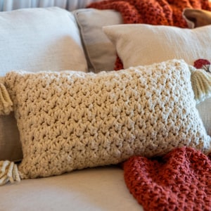 May include: A rectangular cream-colored throw pillow featuring a chunky crochet knit texture. The pillow is accented with four matching cream-colored yarn tassels, one attached to each of the four corners of the rectangular cushion cover.
