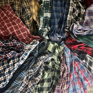 Könnte beinhalten: Ein Haufen karierter Flanellhemden in verschiedenen Farben, darunter Rot, Blau, Grün und Grau. Die Hemden sind gefaltet und gestapelt und zeigen eine Vielzahl von Mustern und Stilen. Das Bild ist aus der Vogelperspektive aufgenommen.