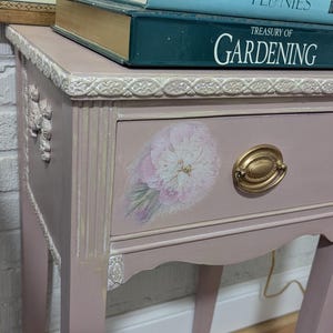 May include: A vintage, pink painted side table with a floral design on the drawer. The drawer has a gold-colored handle. Two books are stacked on top, one titled "Peonies" and the other "Treasury of Gardening".