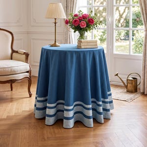 May include: A round blue tablecloth featuring two horizontal light blue stripes near the bottom hem. The fabric drapes in soft folds around the circular table, creating a clean and structured appearance that covers the entire surface down to the floor.