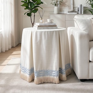 May include: A round side table covered with a floor-length cream-colored fabric cloth featuring box pleats, a light blue embroidered geometric border near the hem, and a thick fringe trim along the bottom edge of the fabric.