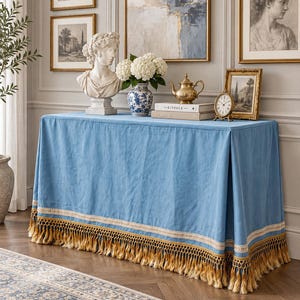 May include: A rectangular table covered with a light blue fabric cloth featuring a cream-colored lace trim and long golden-yellow fringe along the bottom edge. The cloth drapes over the sides of the table, creating a structured, pleated appearance.