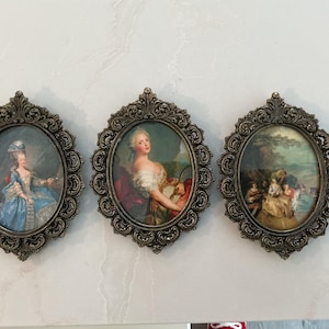 May include: Three antique oval framed prints. The frames have an ornate, dark metallic finish. The prints feature portraits of women in period clothing and a landscape scene. The prints are displayed on a white surface.