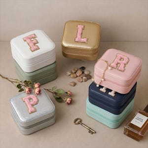 May include: A collection of square jewelry boxes in various colors, each with a letter applique. The boxes are stacked and arranged with a key, perfume bottle, and decorative stones. Colors include white, gold, pink, silver, and blue.