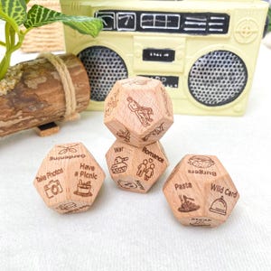 May include: Four wooden, multi-sided dice with engraved images and text. The dice feature illustrations of a camera, a picnic basket, a tank, and a burger. The text includes "Take Photos", "Have a Picnic", "War", "Romance", "Burgers", and "Wild Card".