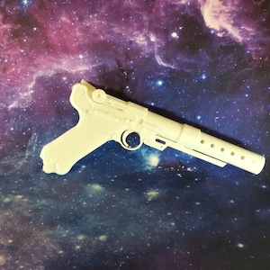 May include: A matte white plastic replica of a Luger pistol featuring a textured grip, a circular trigger guard, and a long barrel with a series of small circular perforations along the side, presented in a side profile view.