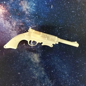 May include: A light-coloured replica revolver pistol, set against a dark blue background with a galaxy-like pattern. The gun features a trigger, hammer, and barrel. The pistol is a detailed model.