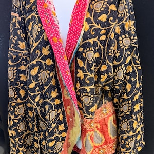 May include: A black jacket with a floral pattern in gold. The jacket's interior features a pink and red patterned fabric. The jacket is open, revealing the lining and a white garment underneath. The jacket is made of a quilted fabric.