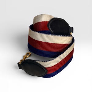 May include: A striped shoulder strap in red, white, and blue. The strap is made of a woven material and has black leather accents at each end. Gold-colored metal clasps are attached to the leather.