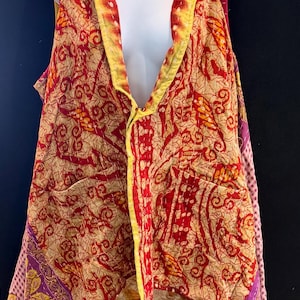 May include: A sleeveless vest with a vibrant, patterned design. The vest features a mix of red, yellow, and beige hues with intricate patterns. It has a draped collar and two front pockets. The edges are trimmed with yellow and pink fabric.