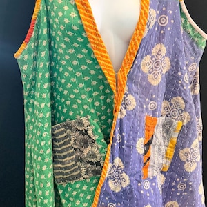 May include: A patchwork vest in shades of green, purple, and orange. The vest features a V-neck and a pocket. The fabric has a quilted texture with floral and geometric patterns. The edges are trimmed with orange fabric.