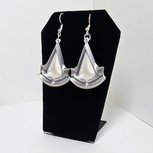May include: A pair of silver-colored earrings with a geometric design. The earrings are displayed on a black velvet jewelry stand against a white background. The earrings have a triangular shape with a decorative edge.