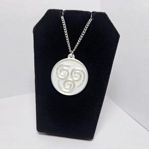 May include: A circular white pendant featuring three embossed spiral designs arranged in a triangular pattern. The pendant is suspended from a silver-toned metal chain, creating a necklace that displays a clean, monochromatic aesthetic.