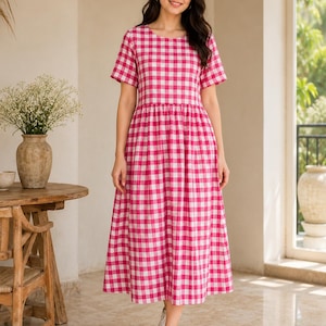 May include: A midi-length dress featuring a vibrant pink and white gingham check pattern, short sleeves, a round neckline, and a gathered waistline that flows into a full, loose skirt. The garment is paired with simple white trainers.