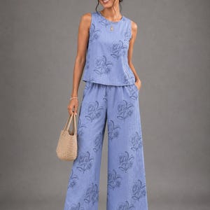 May include: A periwinkle blue sleeveless top and matching wide-leg trousers set featuring a dark blue floral print. The fabric has a subtle vertical ribbed texture. The trousers include side pockets and a relaxed fit, paired with a woven straw bag.