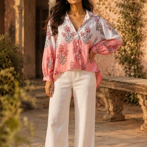 Puede incluir: Una camisa de manga larga con botones con un estampado floral en rojo y gris sobre un degradado blanco y rosa. La camisa se combina con pantalones blancos de pernera ancha. El atuendo es adecuado para ropa informal.