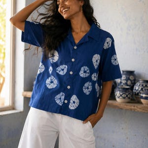 May include: A short-sleeved, indigo blue shirt with white, circular tie-dye patterns. The shirt has a classic collar and white buttons. Paired with white, wide-leg pants. The background features blue walls and decorative pottery.