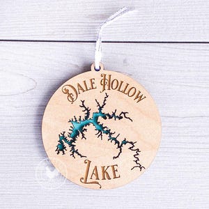 May include: A round wooden ornament with the words "Dale Hollow Lake" engraved in a decorative font. The lake's outline is etched and filled with teal-colored resin. A white ribbon is attached for hanging.