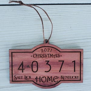 May include: A wooden Christmas ornament with the text "2021 CHRISTMAS 40371 SALT LICK HOME KENTUCKY" engraved in a dark brown color. The ornament has a rectangular shape with a decorative top and a brown string for hanging.