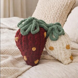 May include: Two strawberry-shaped decorative pillows. One is a deep red with yellow seed details and green leaves, the other is off-white with yellow seeds and green leaves. The pillows are on a bed with neutral-colored bedding.